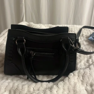 Svart handväska från Gina Tricot - Snygg svart handväska med både handtag och axelrem från Gina Tricot. Väskan har flera fack med dragkedja, detaljerade sömmar och stilren design. Materialet är skinnimitation och väskan har silverfärgade metalldetaljer. Perfekt storlek för det viktigaste.