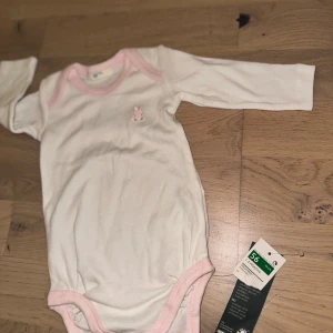 Vit långärmad babybody med rosa kant Eton Et Fils vit, storlek 56 - Vit body med långa ärmar och rosa detaljer runt halsen och benöppningarna. En liten broderad kanin finns på bröstet. Söt vit långärmad body från Newbie i storlek 56, med ljusrosa kant runt hals och ben. Broderad liten kanin på bröstet. Mjuk och skön bomull, perfekt för de allra minsta. Tryckknappar i grenen för enkel på- och avklädning.