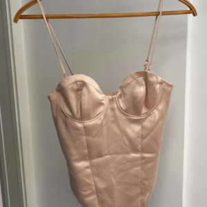 Champagnefärgad satin korsettopp - Snygg korsettopp i champagnefärgad satin med tunna justerbara axelband och markerade kupor. Toppen har en glansig finish och figurnära passform som ger en lyxig känsla. Perfekt att styla med jeans eller kjol för en trendig look. 