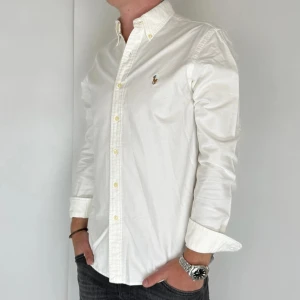 Ralph Lauren Skjorta - Nu har vi nöjet att erbjuda en klassisk skjorta från det välkända varumärket Ralph Lauren. Plagget är i storlek Medium och är i mycket gott skick, utan några synliga defekter. Modellen på bilden är 180 cm lång och väger 75 kg, vilket kan ge en god indikation på tröjans passform.