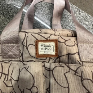 Beige Winnie the Pooh skötväska  - Superfin beige skötväska  med brunt konturmönster av Nalle Puh över hela väskan. Väskan har två handtag, flera fack både inuti och utanpå samt dragkedja upptill. Tillverkad i slitstark bomullscanvas och har en söt etikett framtill.