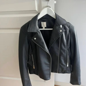 Svart skinnjacka med dragkedjor - Snygg svart skinnjacka med klassisk bikerstil och silvriga dragkedjor. Jackan har breda slag, två framfickor med dragkedja och en croppad passform som ger en cool vibe. Perfekt för dig som vill ha en edgy look till vardags.