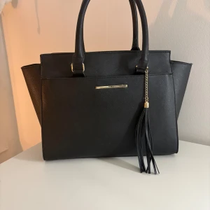 Svart handväska från Anna Field - Stilren svart handväska från Anna Field med guldfärgade detaljer och en snygg tofs i skinnimitation. Väskan har två rejäla handtag och en avtagbar axelrem, perfekt för att bära på olika sätt. Klassisk form med strukturerad yta och metallplatta med logga framtill. Använd ett fåtal gånger men inga defekter💞