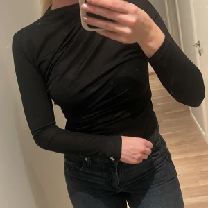 Svart långärmad polotopp - Säljer en svart långärmad polotopp i stretchigt och mjukt material. Toppen har en lätt draperad effekt över bröstet och en smal passform som sitter snyggt mot kroppen. Perfekt att styla med jeans eller kjol för en clean look.