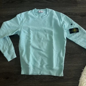 Ljusblå sweatshirt från Stone Island - Säljer en ljusblå sweatshirt från Stone Island med klassisk rund hals och ribbade muddar. Tröjan har den ikoniska loggan på vänster ärm och är tillverkad i mjuk bomull. Perfekt för dig som gillar stilrena och bekväma plagg.