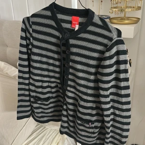 Randig kofta från Esprit med knappar och kurvor vid midjan - Snygg kofta från Esprit i svart och grått med horisontella ränder. Koftan har rund hals, långa ärmar och knäppning med svarta knappar framtill. Två fickor med liten rosa stjärndetalj ger extra stil. Stickad i mjukt material, perfekt för lager på lager.