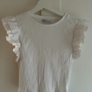 Vit ribbad topp med volangärmar från Zara - Säljer en vit ribbad topp från Zara i storlek L. Toppen har rund hals och ärmlös design med breda, dubbla volangdetaljer på axlarna som ger en extra touch. Materialet är mjukt och stretchigt, perfekt för dig som gillar stilrena och feminina plagg.