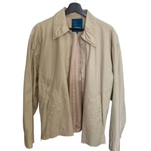 J Lindeberg Overshirt  - Snygg beige vindjacka från Limabhera med klassisk krage, dragkedja framtill och praktiska fickor. Jackan har lång ärm, justerbara detaljer i midjan och en cool ficka med knapp på ryggen. Perfekt för dig som gillar stilren och enkel design.