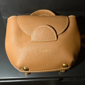 Liten brun crossbody väska från Polène i skinn - Supersnygg brun handväska från Polène i äkta skinn med guldfärgade metalldetaljer. Väskan har ett stilrent, rundat lock, justerbar axelrem och praktiskt handtag.