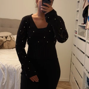Svart stickad omlott-tröja med pärlor - Säljer en svart stickad omlott-tröja med v-ringning och långa ärmar. Tröjan har dekorativa pärlor över hela och ett snyggt hålmönster. Midjan är ribbad för en smickrande passform. Perfekt för dig som vill ha något extra till din outfit.