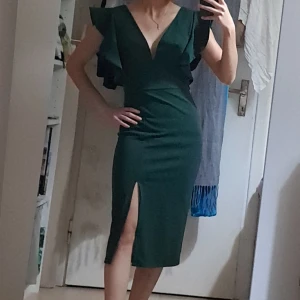 Mörkgrön klänning från WalG - Supersnygg mörkgrön klänning från WalG med v-ringning, volangärmar och slits framtill. Klänningen är figurnära och har en elegant midilängd. Perfekt för dig som vill sticka ut med stilrena detaljer och en djup rygg. Materialet känns mjukt och följsamt. Storlek xs