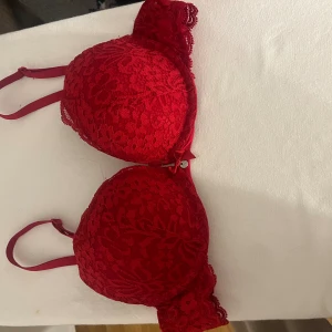 Röd spets-bh från Hunkemöller - Säljer en snygg röd bh från Hunkemöller med blommig spets, vadderade kupor och justerbara axelband. Dekorerad med en liten rosett och metallcharm framtill. Använd fåtal gånger o i princip i ny skick. Ny pris 459kr