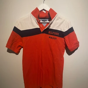 Röd och vit Tommy Hilfiger piké - Snygg pikétröja från Tommy Hilfiger i rött, vitt och svart med broderad logga och romerska siffror på bröstet. Barnstorlek L 16-18 år. Kan bäras som S. 