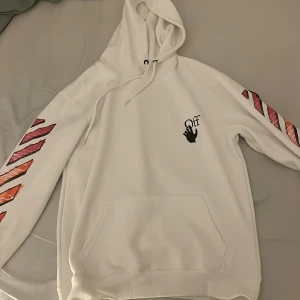 Vit Off-White hoodie med färgglada detaljer - Säljer en vit hoodie från Off-White i storlek S. Tröjan har huva, magficka och Off-White-logga på bröstet. Den har en liten fläck och armarnas pil märken är lite trasiga. Ärmarna och ryggen har ikoniska pilar och ränder i rosa, rött och orange. 