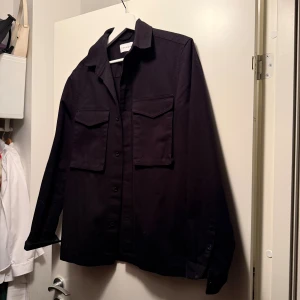 Svart overshirt från Topman - Stilren svart overshirt från Topman med klassisk krage, två stora bröstfickor med lock och knäppning framtill. Skjortan har lång ärm och är tillverkad i ett kraftigare bomullstyg, perfekt för lager-på-lager. Enkel och clean look som funkar till allt. Storlek XS men oversize så passar S som en smäck, jag bär vanligtvis S