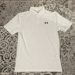 Vit performance polo från Under Armour - Klassisk vit performance polo från Under Armour med korta ärmar och svart logga på bröstet. Skjortan har krage och knappar framtill. Tillverkad i ett lätt och ventilerande syntetmaterial som passar perfekt för sportiga aktiviteter.