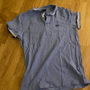 Blå pikétröja från BOSS - Snygg blå pikétröja från BOSS med klassisk krage och knappar framtill. Tröjan har korta ärmar med vit och mörkblå kant, samt diskret BOSS-logga på bröstet. Perfekt för dig som gillar stilrena och sportiga plagg.