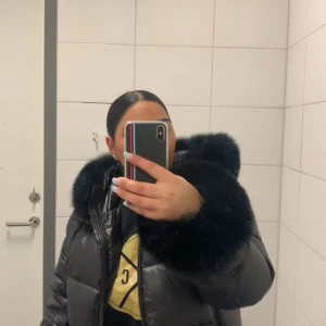 Svart pufferjacka med fuskpäls från Mocekgoo - Säljer en svart pufferjacka från Mocekgoo med stor fluffig fuskpäls runt huvan. Jackan har dragkedja framtill och är riktigt tjock och varm, perfekt för kalla dagar. Klassisk pufferstil med breda kanaler och snygg glansig finish.Går även att använda utan päls 