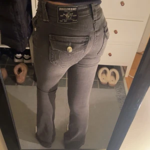 Grå bootcut jeans från True Religion - Snygga grå jeans från True Religion i modellen Becca (färg charcoal) med bootcut passform och mid waist. Jeansen har klassiska fem fickor, dekorativa sömmar och en ikonisk bakficka med knapp. Materialet är mjukt jeans-tyg med stretch för skön passform. Endast använda en gång. (Jag är 165)