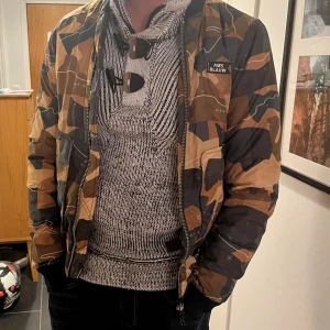  bomberjacka Scotch & Soda - Säljer en snygg bomberjacka från Scotch & Soda med camouflagemönster i brunt, beige och svart. Jackan har dragkedja framtill, ribbade muddar och orange foder. Märket 'AMS BLAUW' syns på bröstet. Skriv till mig vid frågor eller pris! Inga defekter 