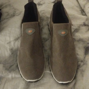 Bruna slip-on boots från ZOOO - Säljer ett par bruna slip-on boots med robust svart sula och orange detaljer. Skorna har en rund tå och är tillverkade i mocka med elastiska paneler på sidorna för enkel påtagning. Perfekta för dig som gillar en stilren och sportig look. Dom är helt nya aldrig använda. 