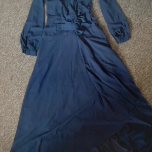 Marinblå långklänning med knappar - Snygg marinblå långklänning med långa ärmar och markerad midja. Klänningen har omlottkänsla framtill och fina knappar vid ärmsluten. Tillverkad i ett mjukt, lätt material som faller snyggt. Perfekt för dig som gillar stilrena och eleganta plagg. Aldrig använd!