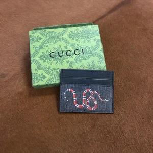 Gucci plånbok med ormmotiv - Snygg svart plånbok från Gucci med ett coolt ormmotiv i rött, vitt och svart på framsidan. Plånboken har flera kortfack och är tillverkad i slitstarkt skinn med diskret Gucci-mönster i bakgrunden. Perfekt för dig som gillar statement-accessoarer.