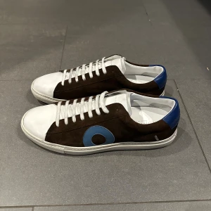 Oliver Cabell sneakers brun/blå/vit - Snygga sneakers från Oliver Cabell i brunt mocka med vita skinnpartier och blå detaljer på häl och sida. Klassisk låg modell med vit platt sula och snörning. Oanvända men lite smutsiga då de legat på ett lager 