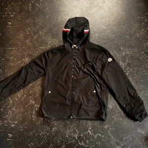 Svart vindjacka från Moncler med huva - Svart vindjacka från Moncler med huva och dragkedja. Jackan har Moncler-logga på ärmen och en snygg rödvit rand på huvan. Tillverkad i lätt polyester, perfekt för blåsiga dagar. Jackan har även NFC skann, skriv för fler bilder
