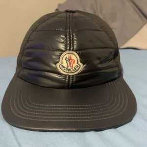 Svart Moncler keps i quiltad stil - Snygg svart keps från Moncler med quiltad front och klassisk böjd skärm. Kepsen har Monclers ikoniska logga framtill och är tillverkad i en blandning av syntetmaterial och bomull. Perfekt accessoar för en stilren och sportig look.