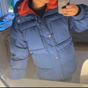 Blå pufferjacka med orange foder - Säljer en blå pufferjacka med huva och orange foder. Jackan har stora fickor framtill, ribbade muddar vid ärmslut och stängs med dragkedja och knappar. Perfekt för kalla dagar och har en loose passform som ger skön streetkänsla. Använd under en vinter
