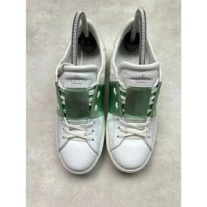 Valentino Garavani sneakers med grön detalj - Snygga vita sneakers från Valentino Garavani med en metallicgrön bred rem över vristen. Skorna har klassisk snörning, platt sula och coola nitar baktill. Tillverkade i skinn och har en modern, stilren look som sticker ut. Strl 38