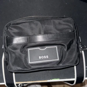 axelväska från BOSS/ It diskussion - Snygg svart axelväska från BOSS med stilren design och logga framtill. Väskan har flera fack med dragkedja och justerbar axelrem med vit detalj. Tillverkad i slitstark polyester med detaljer i skinnimitation. Perfekt för att bära dina viktigaste grejer.