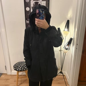 Svart parkajacka från Bondelid - Svart parkajacka från Bondelid med huva och snörning. Insidan är fodrad med mjuk teddyfleece i vitt och har röda detaljer längs kanten. Jackan har praktiska fickor och dragkedja framtill, perfekt för kyliga dagar.