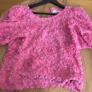 Rosa blommig blus från Holly & White - Superfin rosa blus från Holly & White med 3D-blommor och spetsdetaljer över hela toppen. Blusen har korta puffärmar och rund halsringning. Perfekt för dig som vill sticka ut med en romantisk och färgglad look.