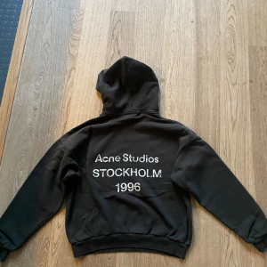 Svart hoodie Acne Studios Stockholm - Svart hoodie från Acne Studios med stor vit text på ryggen: 'Acne Studios STOCKHOLM 1996'. Klassisk passform . Perfekt för dig som gillar streetstyle och vill ha en stilren look med tydlig logga.