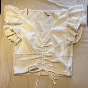 Vit blus med volangärmar - Supersöt vit blus i storlek XS. Blusen har volangiga korta ärmar, v-ringning och snygg snörning framtill med knyt. Materialet är lätt och har broderade hålmönster som ger en fräsch vibe. Perfekt för dig som gillar romantisk och feminin stil.