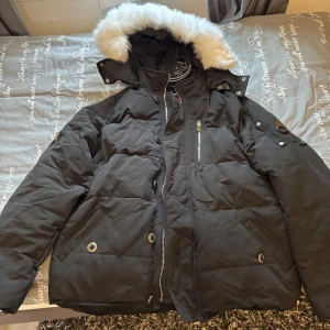 Svart dunjacka Moose Knuckles - Svart dunjacka från Moose Knuckles med fluffig vit fuskpäls på huvan, coolt metallmärke på ärmen och flera fickor med dragkedja och knappar. Helt ny! 