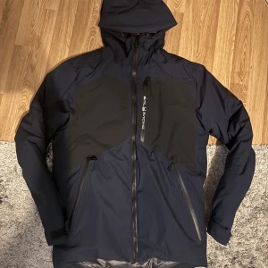 Mörkblå vinterjacka  från Sail Racing - Snygg mörkblå och svart vindjacka från Sail Racing med huva och flera dragkedjor. Jackan har en sportig look med logga på bröstet och rygg, samt praktiska fickor med dragkedja. Perfekt för dig som gillar stilren och funktionell design.