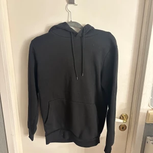 Svart hoodie XS med huva och ficka - Svart hoodie i storlek XS med klassisk huva och snörning. Tröjan har en stor magficka framtill och är långärmad. Perfekt för chill dagar och enkel att matcha med jeans eller mjukisbyxor. Materialet känns mjukt och skönt mot huden.