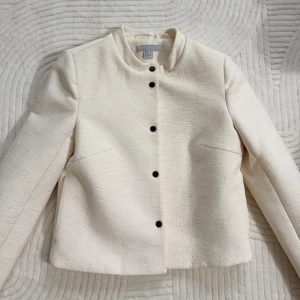 Vit kavajjacka från H&M med struktur - Snygg vit kavajjacka från H&M i storlek 36. Jackan har en cool strukturerad yta, rund krage och svarta knappar framtill. Passformen är rak och den är långärmad, perfekt för att lyfta din outfit med en clean och modern vibe.