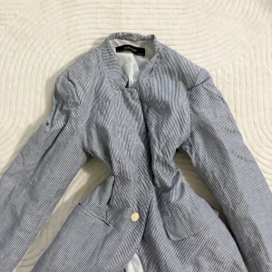 Randig kavaj från Zara Basic - Snygg kavaj från Zara Basic i blått och vitt med smala ränder. Modellen har lång ärm, en knapp framtill och två fickor. Materialet känns lätt och fräscht, perfekt för dig som gillar stilrena och klassiska vibbar.