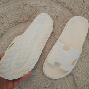 Vita slip-in sandaler med struktur - Säljer ett par vita sandaler i slip-in modell med bred rem och strukturerad yta. De har öppen tå och platt sula med mönstrad undersida för extra grepp. Perfekta för sommaren och riktigt sköna att glida i hemma eller på stranden.