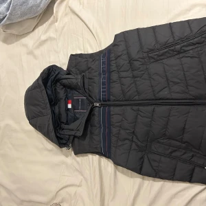 Svart dunväst från Tommy Hilfiger - Svart quiltad dunväst från Tommy Hilfiger med huva och dragkedja framtill. Västen har ett mörkblått band med röd och vit detalj samt Hilfiger-logga över bröstet. Praktiska sidofickor med dragkedja och sportig look.