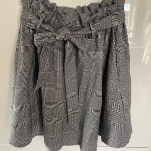 Rutig kjol med knyt från H&M - Superfin rutig kjol från H&M i grått och svart med inslag av vitt och rött. Kjolen har hög midja, papperspåse-linning och ett brett knytband framtill. Modellen är kort och har en lös passform som ger en chill vibe.