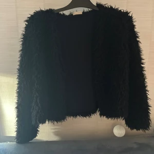 Svart pälsjacka i fuskpäls barn  - Säljer en svart pälsjacka i fuskpäls med långärmad design. Jackan har en fluffig och mjuk känsla, perfekt för att ge en cool och trendig look. Passformen är normal och jackan är helsvart utan synliga detaljer eller mönster.