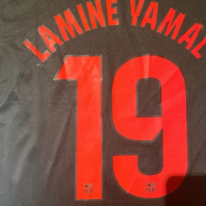 FC Barcelona x Cactus Jack fotbollströja - Snygg svart FC Barcelona fotbollströja med röd nummer 19 och Lamine Yamal på ryggen. Framsidan har silverfärgad Nike-logga, svart FCB-emblem med röd och blå kant, samt Cactus Jack-grafik. Materialet är lätt och andas, perfekt för match eller träning.