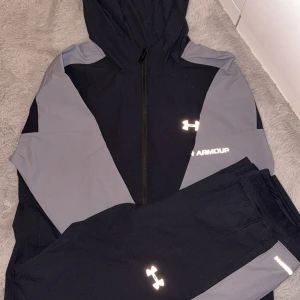 Svart och grå hoodie från Under Armour - Snygg svart och grå hoodie från Under Armour med dragkedja och huva. Tröjan har långa ärmar och coola logodetaljer på bröstet. Perfekt för träning eller chill, med sportig look och bekvämt material som andas.             Finns i fler färger mörkblå/grå, ljusgrå/ svart och den på bilden. Finns alla storlekar från s till xl.       Bara att höra av sig för bilder.