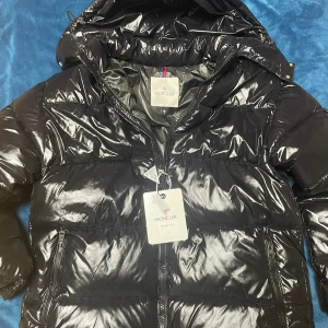 Moncler Maya - Säljer en svart, glansig pufferjacka från Moncler i storlek L. Jackan har huva, två sidofickor med dragkedja. Perfekt för dig som vill ha en varm och snygg jacka med street vibe och exklusiv känsla.