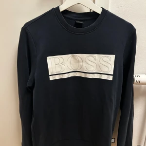 Mörkblå långärmad tröja Hugo Boss - Mörkblå långärmad tröja från Hugo Boss med stor vit logotyp på bröstet. Rund hals och mjukt material, perfekt för en clean och stilren look. Passar dig som gillar klassiska märken och enkel design.